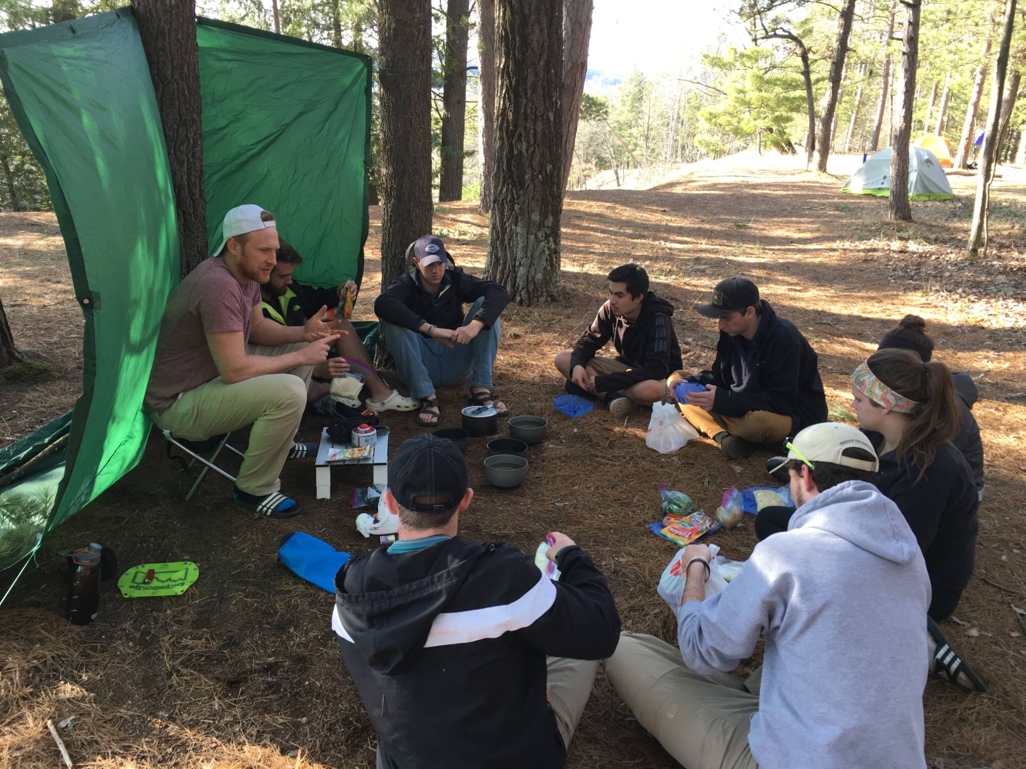 students and instructor sitting on the ground of a shady forest having a lesson on camp cooking
